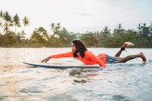 Black Long-haired Man Paddling On Long Surfboard To The Surfing Spot In Indian Ocean. Palm Grove Litted Sunset Rays In The Background. Extreme Water Sports And Traveling To Exotic Countries Concept.