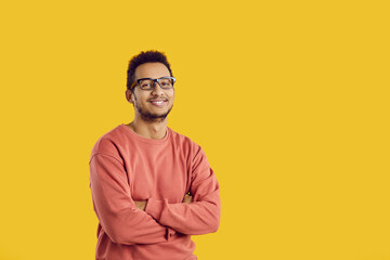 Studio portrait of happy male university student in glasses. Smiling young black man in spectacles standing with his arms crossed isolated on copyspace yellow background with copy space on right side