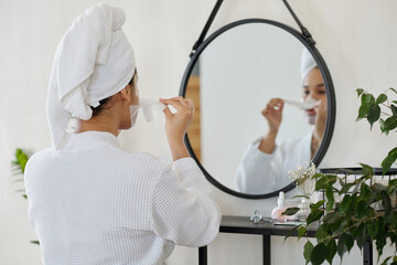 Back of young woman in white bathrobe and with towel on head taking off sheet mask in front of mirror while standing in bathroom