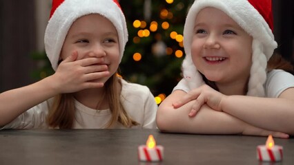 Wall Mural - Happy girls in red santa hat laugh sitting at table on background of Christmas tree at home. Little children spend New Years Christmas holiday time.