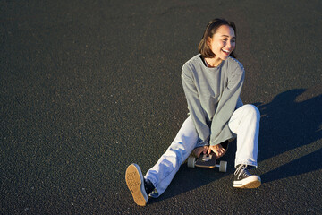 Happy beautiful korean teen girl sits on her skateboard, cruising on longboard, wearing casual clothes