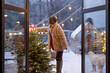 Young woman decorates Christmas tree, attaching festive bow on top while standing on step ladder at beautiful snowy backyard, view through the window. Concept of preparation for the winter holidays