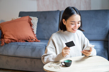 Smiling cute asian woman using credit card and smartphone, paying bills online, holding mobile phone, looking at camera