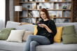 © sofiko14 - Attractive caucasian red hair woman sitting on couch and using modern smartphone indoors. Female freelancer taking break during working process at home.