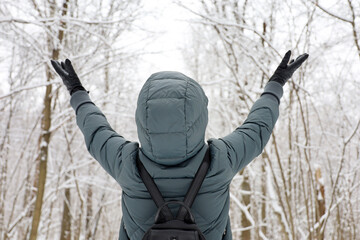 Woman wearing down jacket with hood standing with her hands raised and enjoying the snowy weather. Leisure in winter park, cold season