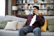 © sofiko14 - Handsome bearded positive man sitting on couch and calling to his friend using smartphone indoors. Smiling male freelancer taking break during working process at home.
