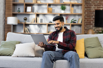 Bearded young man using laptop, sitting on couch at home, handsome guy having video call online in social network, having fun, watching movie, freelancer working on computer project