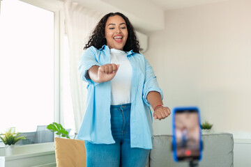 Wall Mural - Cheerful black chubby woman dancing at cellphone camera filming video using phone on tripod at home