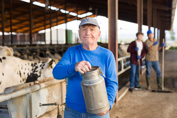 Wall Mural - Positive elderly farmer working at dairy farm, carrying small milk can on background of stall with cows ..