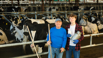 Elderly farmer and his assistant grandson with bottle of milk on the background of cows in a stall