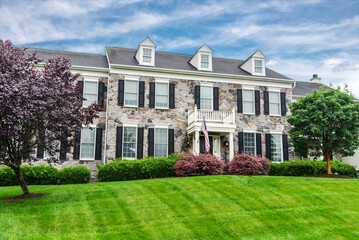 Large modern colonial style house with a balcony. Single family home with large lawn and beautiful landscaping. Sunny day with blue sky.