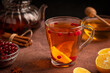 Homemade healthy sweet hot tea drink served in transparent glass cup with red ripe cranberries and lemon citrus fruit slices on dark brown table with teapot, cinnamon spice and honey ingredients