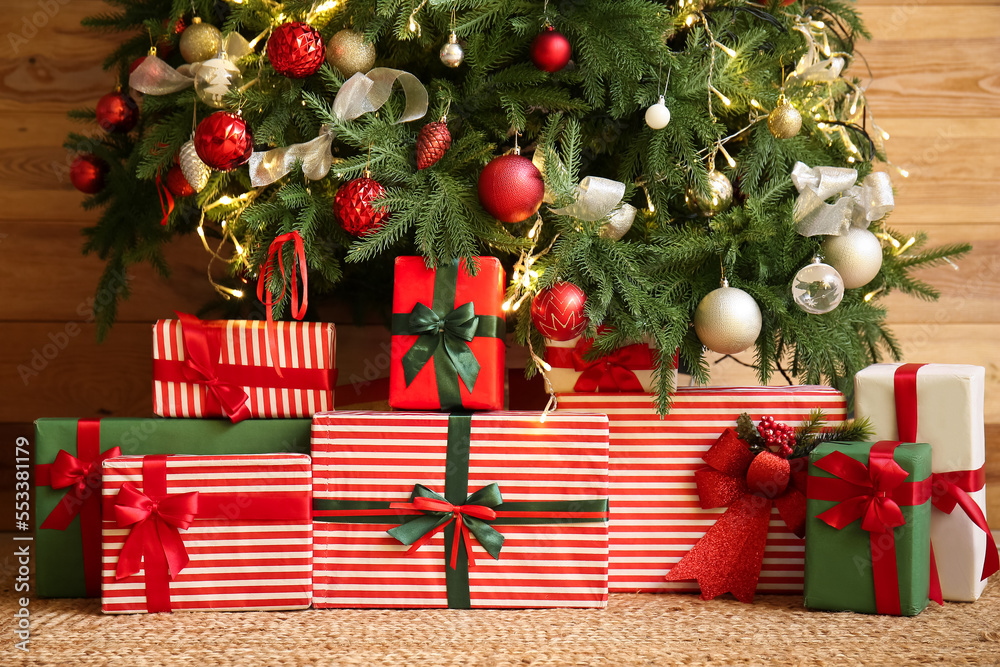 Gifts under glowing Christmas tree near wooden wall
