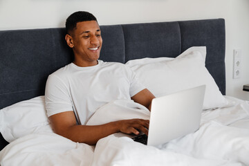 Canvas Print - Joyful african man using laptop while lying in bed at home