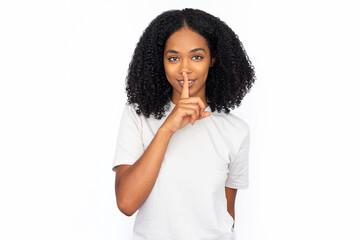 Wall Mural - Pleased multiracial woman pressing finger to lips. Portrait of happy young female model with dark curly hair in white T-shirt looking at camera, smiling and shushing. Silence, secret concept