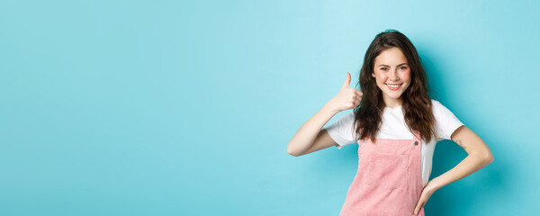 Wall Mural - Portrait of confident and positive young woman show thumb up, say yes, give permission, approve and agree something good, praise nice choice, standing against blue background