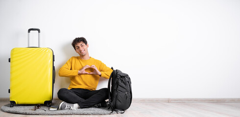 Wall Mural - Pretty romantic young traveler man with suitcase making a heart gesture with her fingers in front of her chest showing her love and affection with a happy tender smile