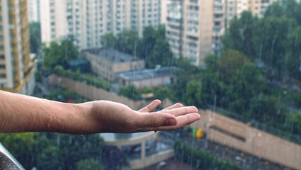 Human hand of an young man under rain.  Raindrops slowly falls onto human palm, closeup, big buildings background. Human feels happy while water drops falling on his hand, in the summer.