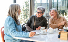 Group Of Elderly Women Have Breakfast In A Cafeteria, Three Retired Female Friends Are Celebrating An Anniversary Drinking Tea And Coffee And Eating Chocolate Cakes