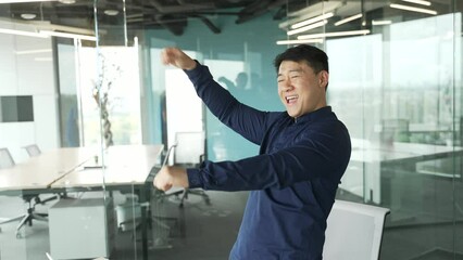 Happy energetic asian businessman IT programmer celebrate success dancing on light glass office in slow motion indoors Smiling cheerful entrepreneur relaxing and having fun alone at modern workplace 