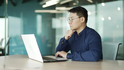 Busy middle aged IT specialist programmer with glasses working typing coding on laptop computer in modern office Asian entrepreneur professional developer thinking analyses finance digital data indoor