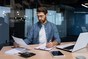 Serious and focused financier accountant on paper work inside office, mature man using calculator and laptop for calculating reports and summarizing accounts, businessman at work in casual clothes.