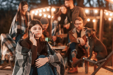 Wall Mural - Woman with smartphone is sitting. Group of people is spending time together on the backyard at evening time