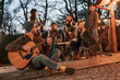© standret - Beautiful man in hat is sitting with his guitar. Group of people is spending time together on the backyard at evening time
