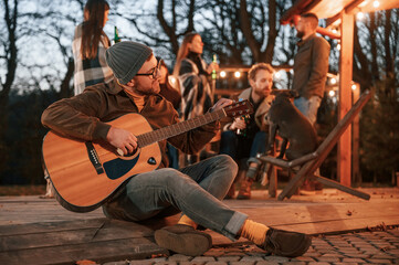 Wall Mural - Beautiful man in hat is sitting with his guitar. Group of people is spending time together on the backyard at evening time