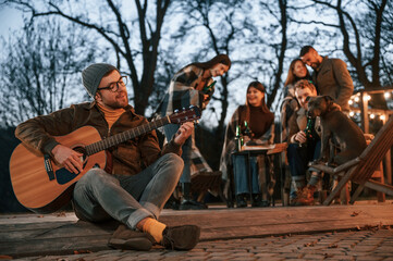 Wall Mural - Man with guitar is sitting in front of his friends. Group of people is spending time together on the backyard