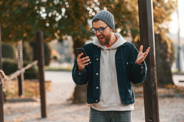 Wall Mural - Pleasantly surprised. With smartphone. Beautiful man in warm clothes is outdoors in the park