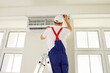 © Studio Romantic - Professional technician worker in uniform and white cap standing on stepleader is checking up, testing, repairing air conditioner indoors, back view. Repair maintenance of electric appliances concept.