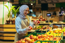 Female Seller In Hijab Browsing And Checking Apples In Supermarket, Woman In Apron Smiling At Work In Store In Fruit And Vegetable Department.