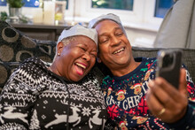 Senior Couple Sitting On Sofa Wearing Christmas Decorations And Using Phone