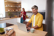 © puhhha - Senior Man Using Laptop At Kitchen. Happy Couple Spending Weekend Together At Home. Blonde Woman Looking At Smartphone At The Background