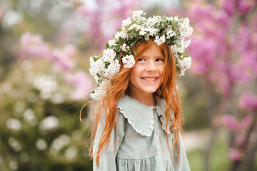 Cute laughing little kid girl 3-4 year old with long curly red hair wear floral wreath and stylish rustic dress over nature background in garden outdoor. Springtime.