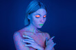 nude woman in bright neon makeup and glowing body paint covering breast with hands isolated on dark blue.