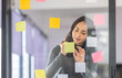 © David - Business female employee with many conflicting priorities arranging sticky notes commenting and brainstorming on work priorities colleague in a modern office.
