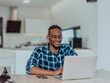 © .shock - African American man in glasses sitting at a table in a modern living room, using a laptop for business video chat, conversation with friends and entertainment