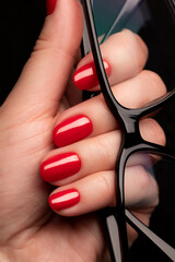 Wall Mural - Female hand with beautiful manicure - red nails with glasses on black background closeup