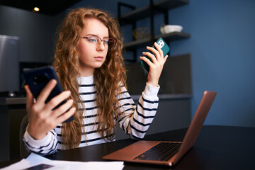 Curly blonde woman in glasses working from home with multiple electronic internet devices. Freelancer businesswoman has tablet, cellphone in hands and laptop on table. Multitasking theme.