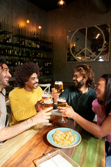 Vertical portrait of a A group of colleague workers toast with beer in the restaurant bar after work at the pub happy hour. Happy friends having fun together drinking alcohol indoors.