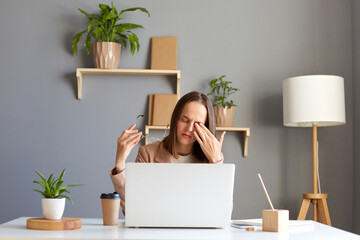 Indoor shot of tired woman wearing official style jacket sitting in office at desk in front of laptop, rubbing her eyes, feels pain, looking at computer display long hours.