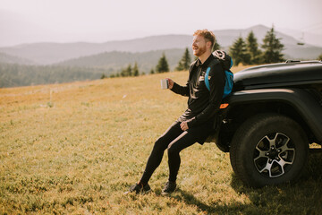 Wall Mural - Young man relaxing and drinking coffee by the terrain vehicle hood at countryside