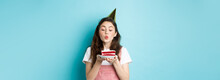 Holidays And Celebration. Excited Woman Celebrating Birthday, Blowing Candle On Cake, Wearing Party Cake And Having Fun, Standing Over Blue Background