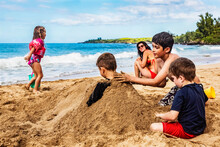Mother Sitting On D. T. Fleming Beach At The Water's Edge And Watching Her Children Playing And Burying A Sibling In The Sand; Kapalua, Maui, Hawaii, United States Of America