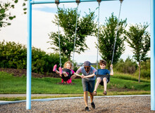 A Father Pushes A Young Girl And Boy On Swings On A Swing Set At A Playground; St. Albert, Alberta, Canada