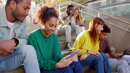 Wall Mural - Happy diverse student friends laughing together while watching funny videos on smart phone sitting outdoors. Young multiracial people having fun using social media mobile phone app. 