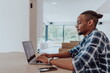 © .shock - African American man in glasses sitting at a table in a modern living room, using a laptop for business video chat, conversation with friends and entertainment