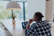 © .shock - African American man in glasses sitting at a table in a modern living room, using a laptop and smartphone for business video chat, conversation with friends and entertainment
